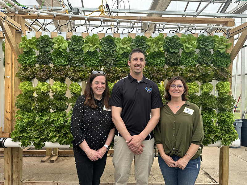 three people smiling, standing in front of a hydroponic garden of greens